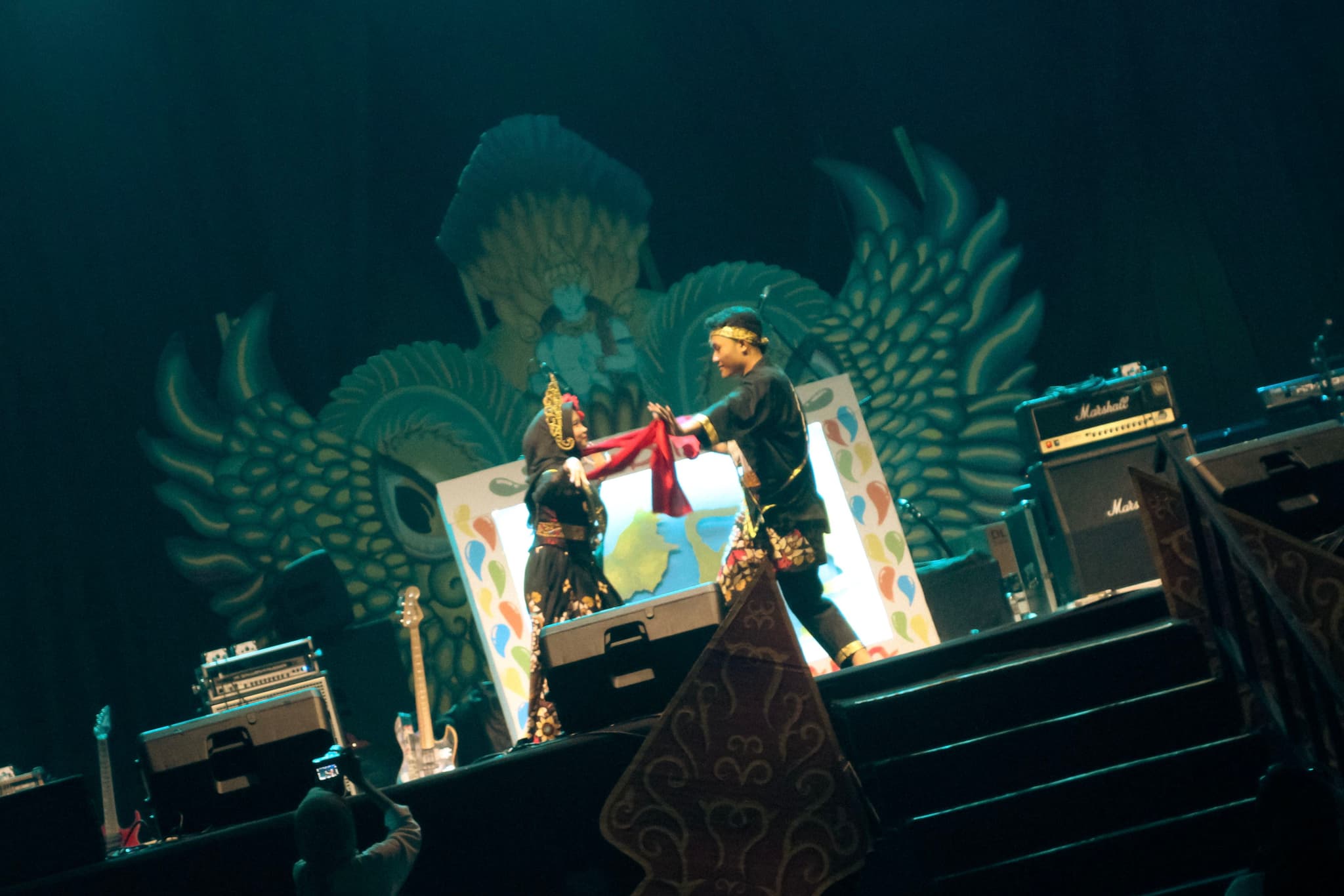 Two performers on stage, one handing a red item to the other, with musical equipment and a large decorative backdrop featuring a bird design