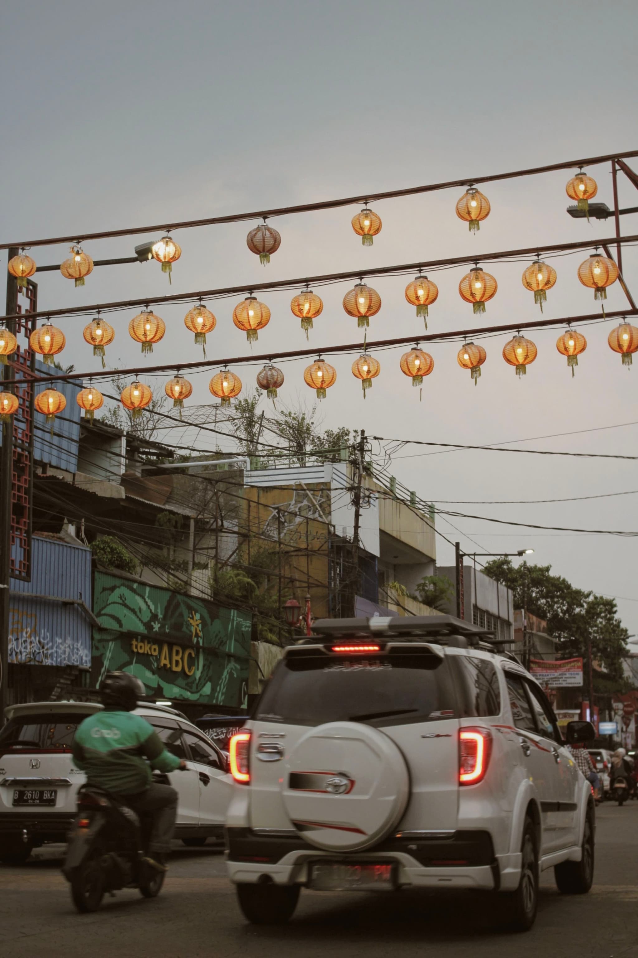 A street scene with vehicles, including a white SUV and a motorbike, under hanging lanterns. Buildings and power lines are visible in the background