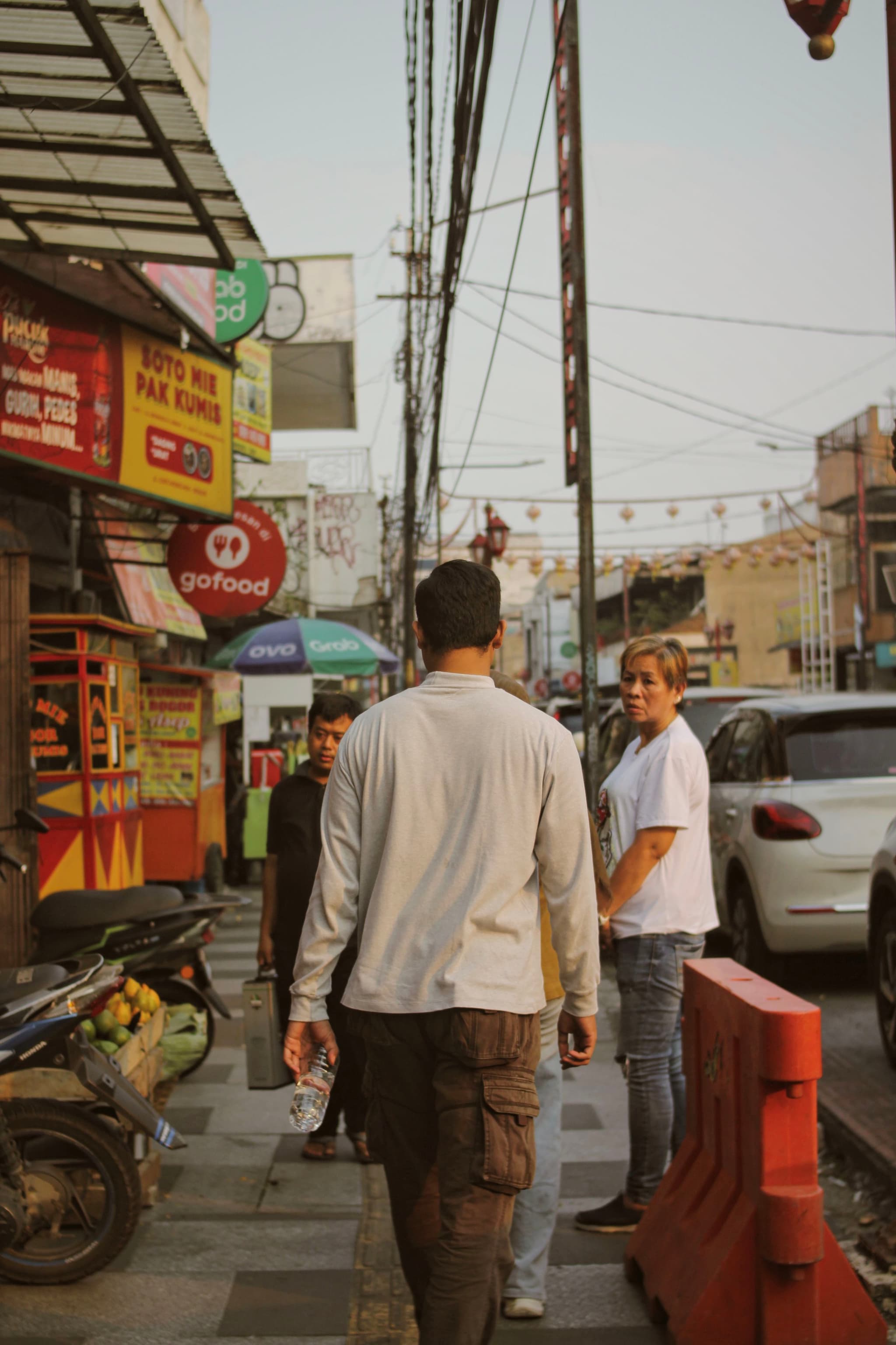 A busy street scene with people walking on the sidewalk, surrounded by shops and parked vehicles. A man in a white shirt is looking back, while another person walks ahead. Bright signage and a motorcycle are visible