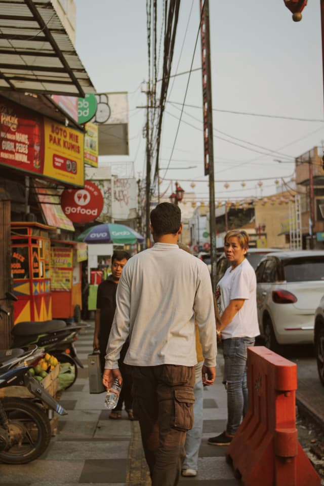 A busy street scene with people walking on the sidewalk, surrounded by shops and parked vehicles. A man in a white shirt is looking back, while another person walks ahead. Bright signage and a motorcycle are visible