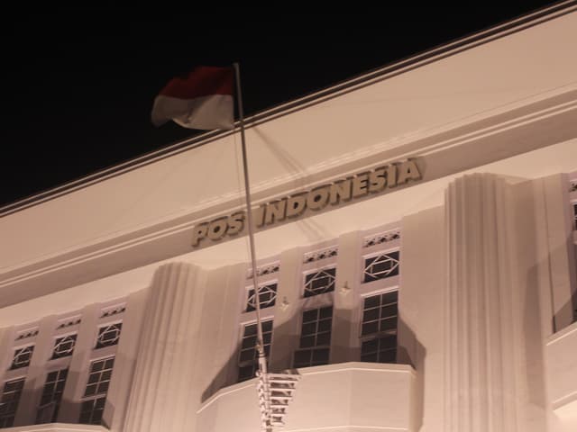 A building with POS INDONESIA on the facade, featuring an Indonesian flag on a pole, illuminated at night