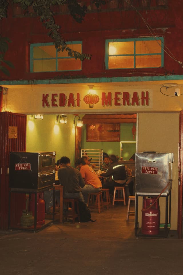 A small, warmly lit eatery named Kedai Merah with people sitting inside, featuring a red and yellow color scheme and a vending machine outside