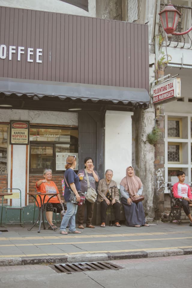 A group of people are sitting and standing outside a coffee shop on a sidewalk. The shop has a brown awning, and there are signs and windows in the background