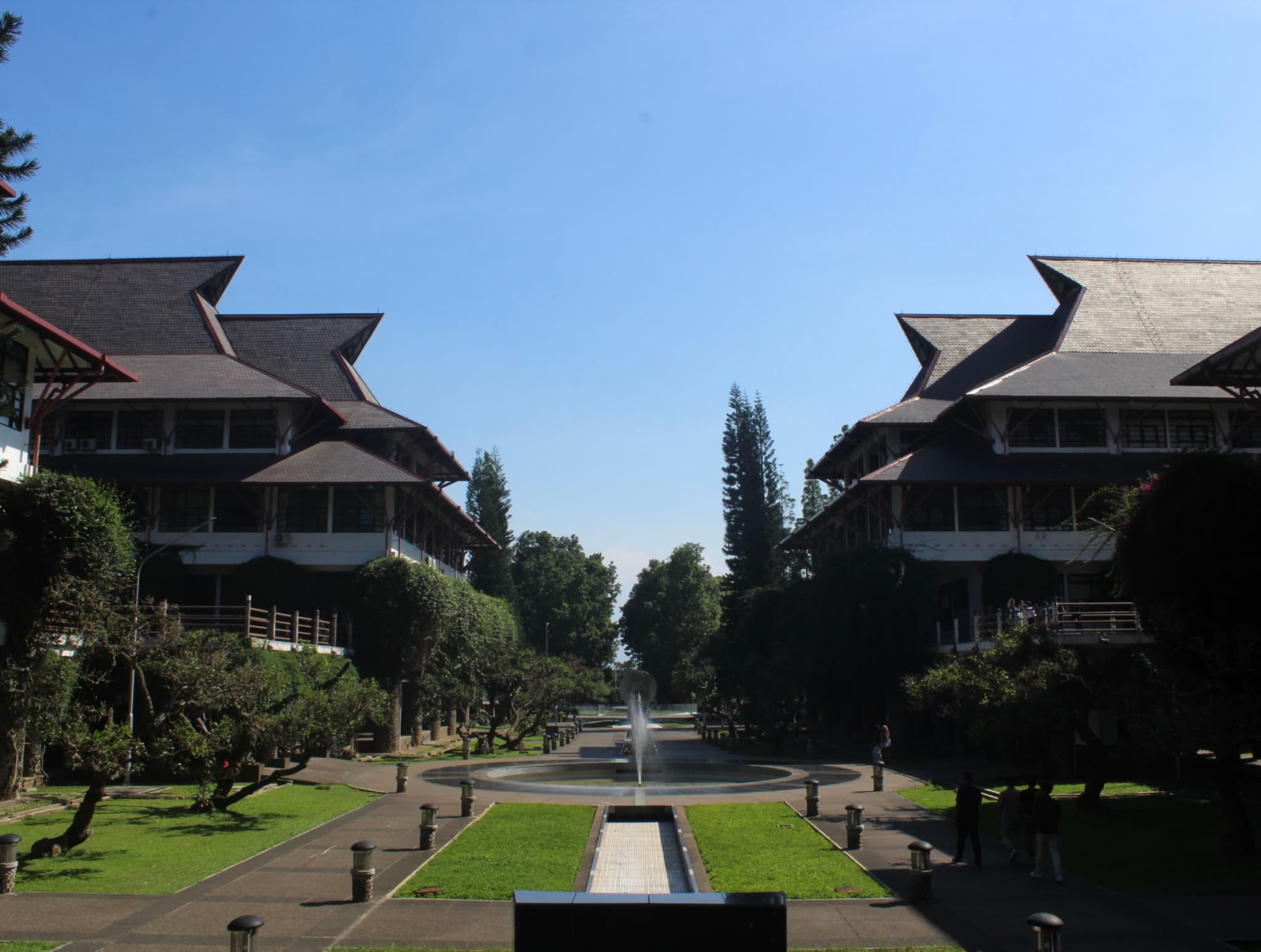 A symmetrical view of two large, traditional-style buildings with tiered roofs, flanking a central pathway and fountain, surrounded by manicured lawns and trees under a clear blue sky