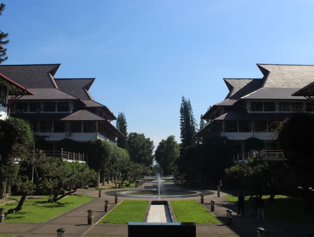 A symmetrical view of two large, traditional-style buildings with tiered roofs, flanking a central pathway and fountain, surrounded by manicured lawns and trees under a clear blue sky