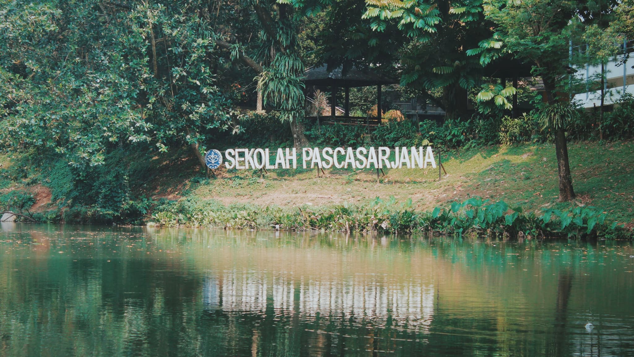 A serene lakeside scene with lush greenery and a sign reading SEKOLAH PASCASARJANA in the background
