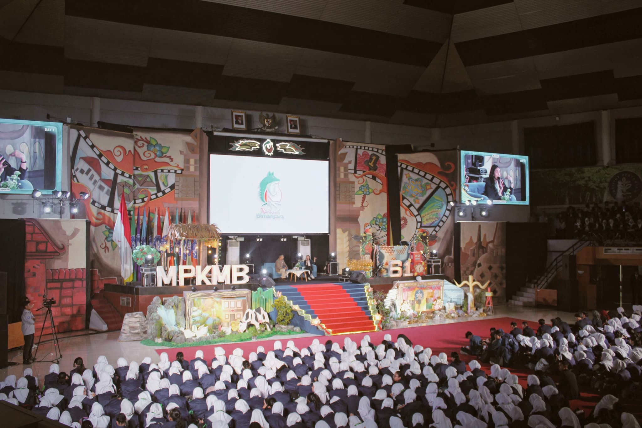 A large indoor event with a decorated stage, featuring a central screen displaying a presentation. The audience, dressed in white, is seated in rows facing the stage. Colorful banners and screens flank the stage, adding to the vibrant atmosphere