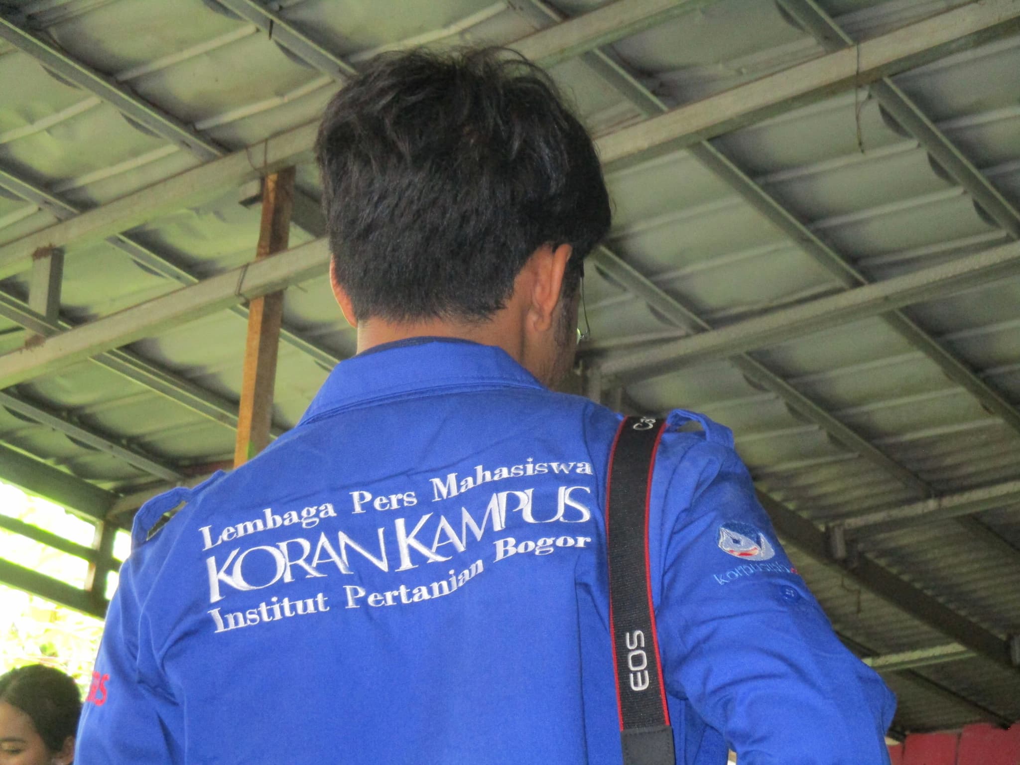 A person wearing a blue jacket with text on the back, standing under a metal roof